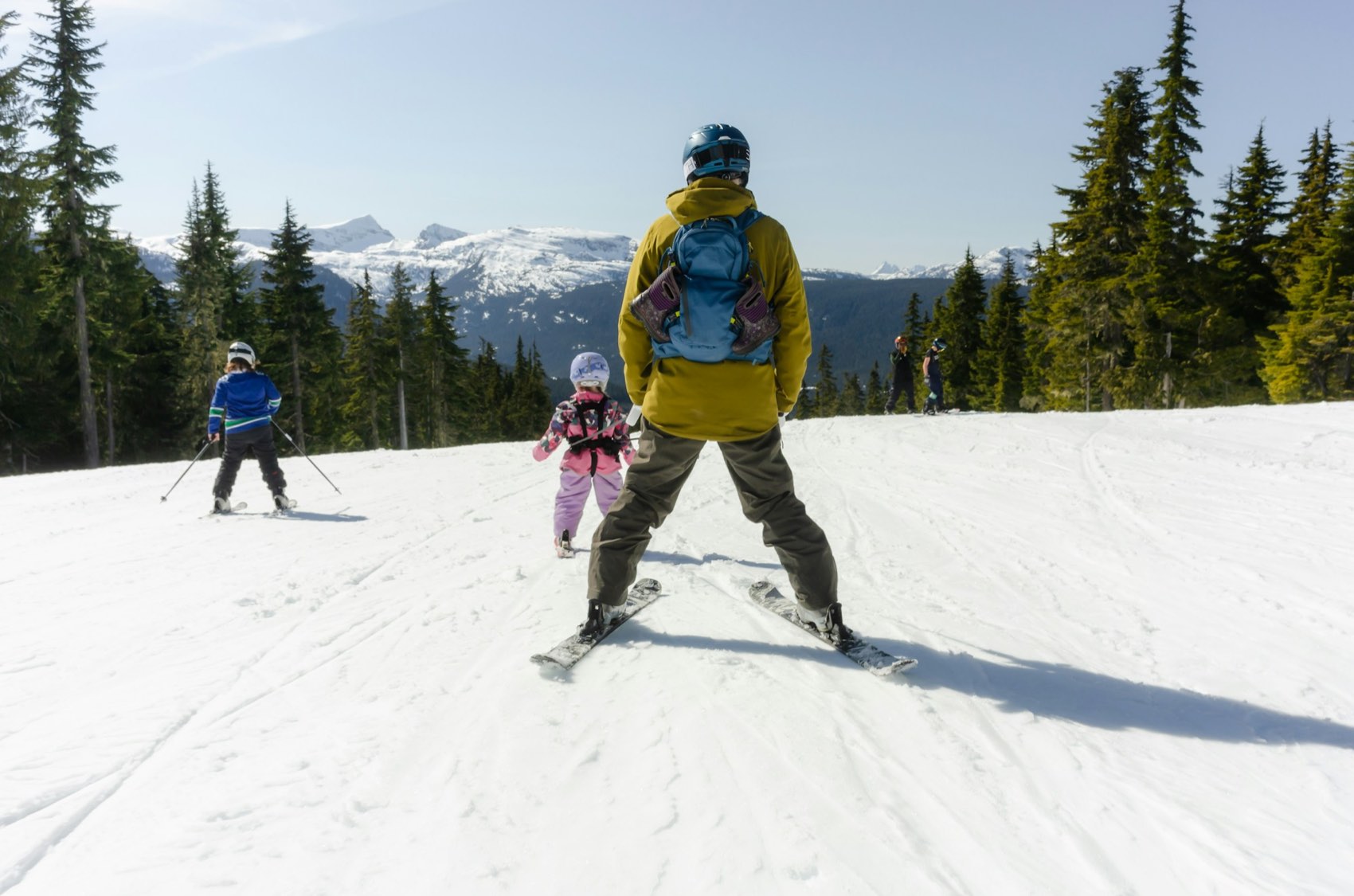 A parent and two children skiing a wide groomed run with evergreen forest and snowy peaks behind