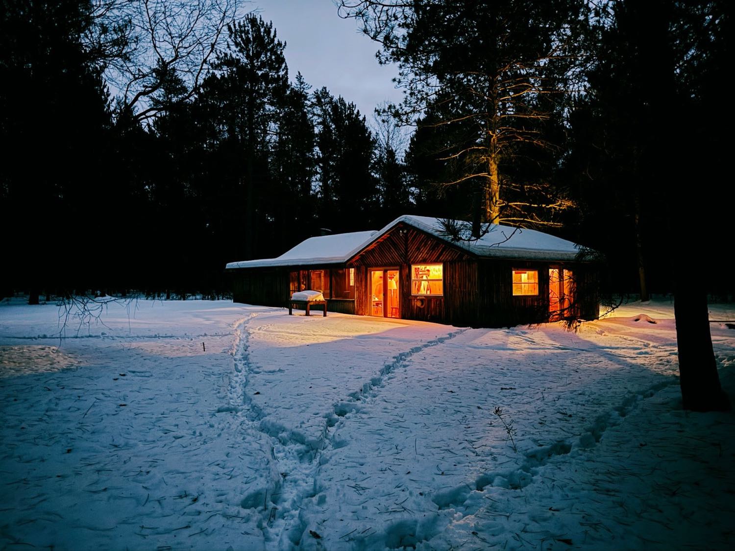 A low timber lodge with warmly lit windows sitting in deep snow among tall pines at dusk