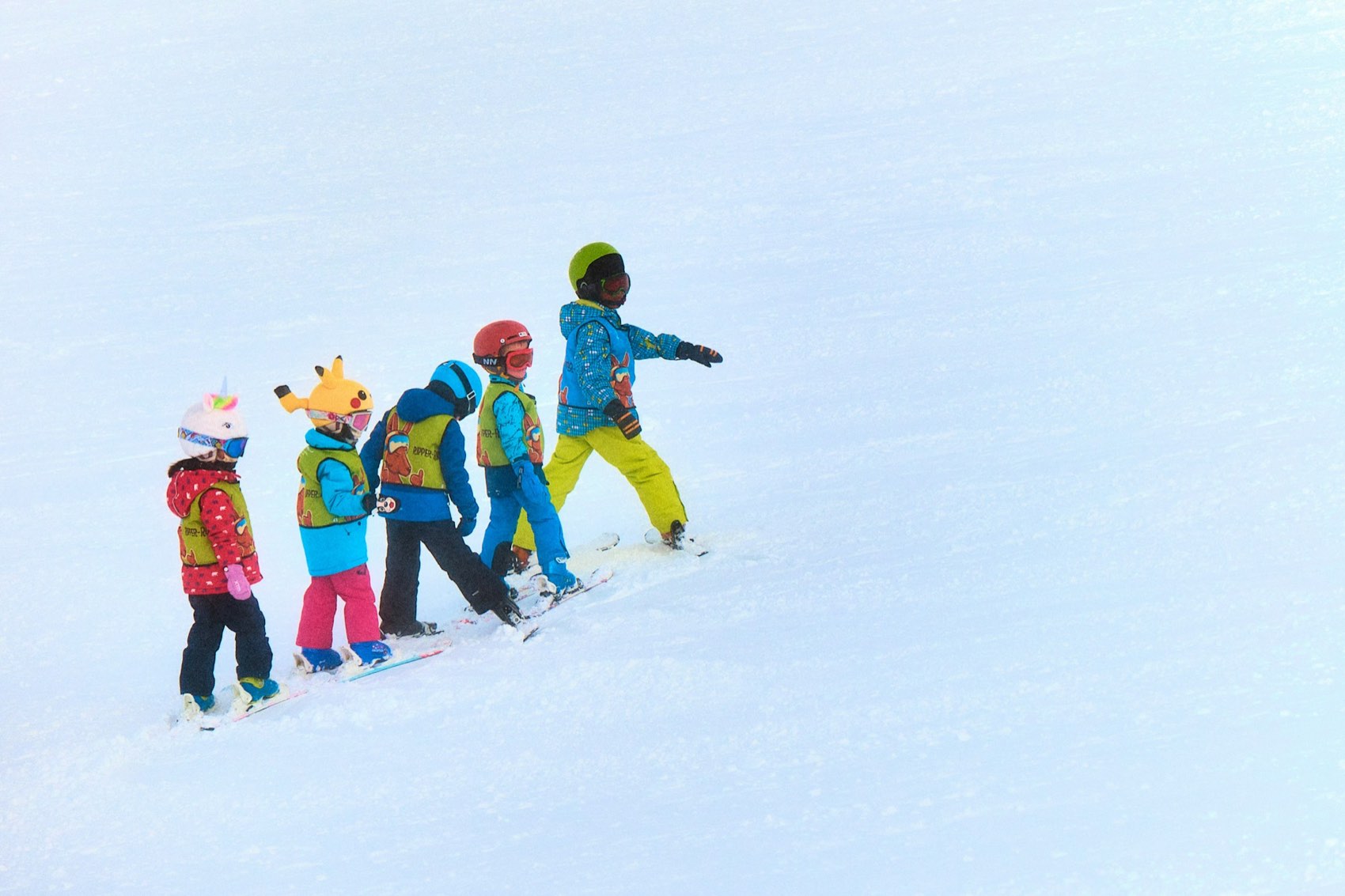 Five small children in colourful helmets and ski-school bibs walking in a line across the snow